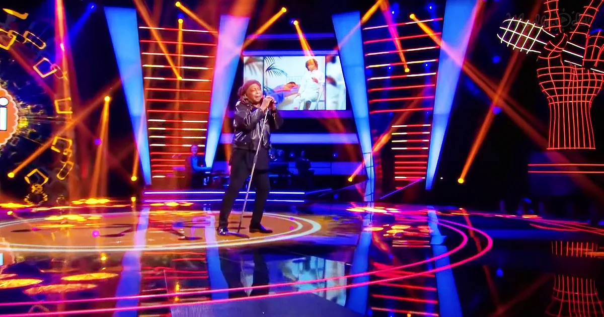 A person in a black outfit sings on a brightly lit stage with colorful lights and a reflective floor.