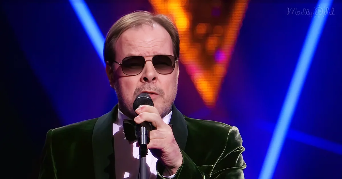 A man in a green suit and sunglasses holds a microphone, singing on stage with blue and orange lights.