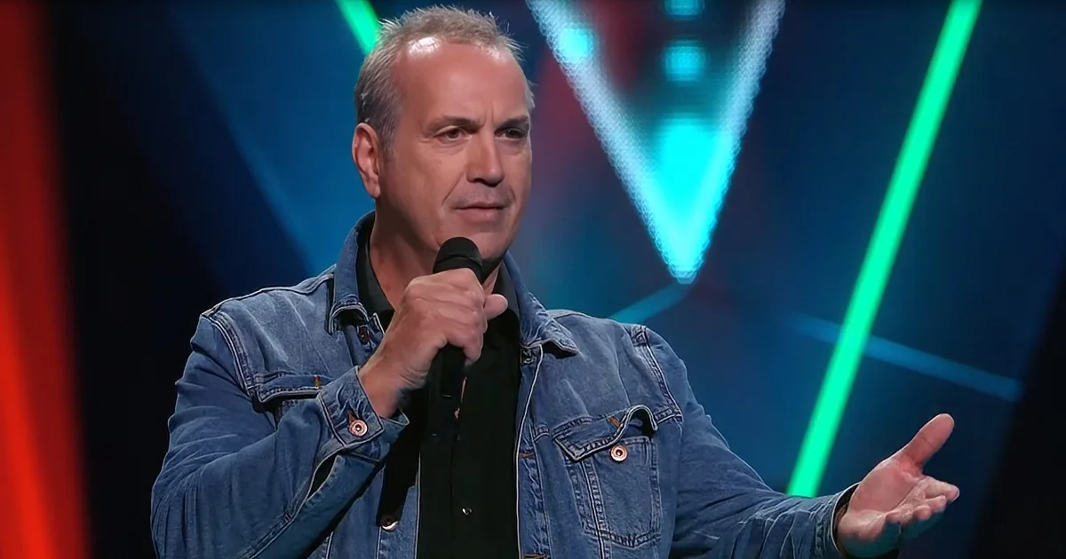 A man in a denim jacket holds a microphone, gesturing with his other hand on stage.