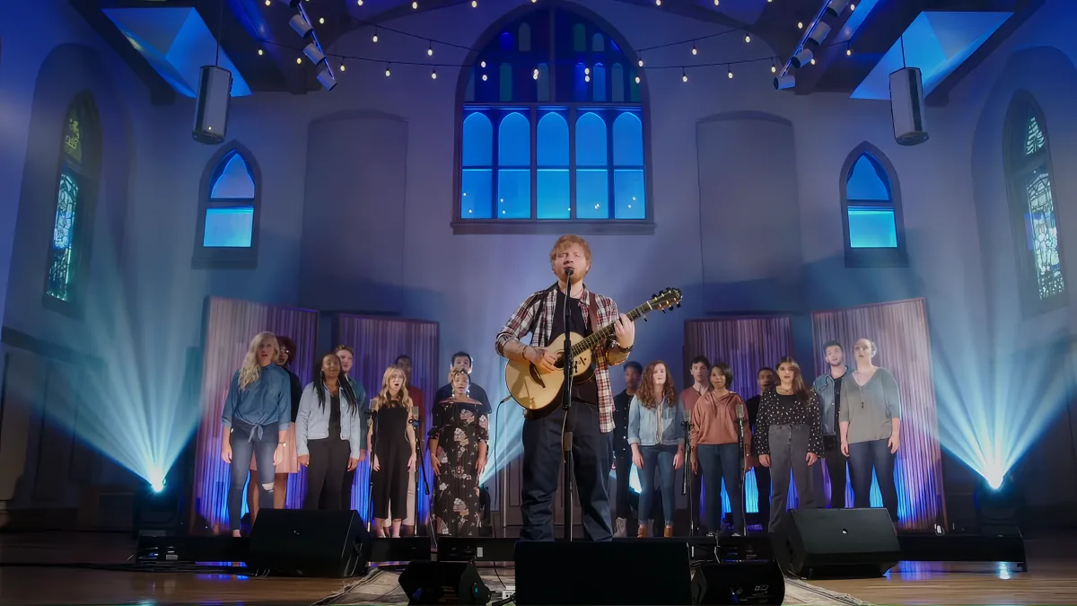 Ed Sheeran plays guitar on stage with a choir in a church setting.