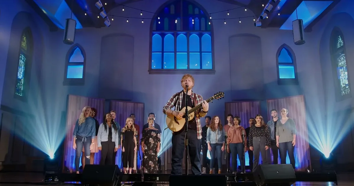 A man plays guitar on stage with a choir in a church setting.