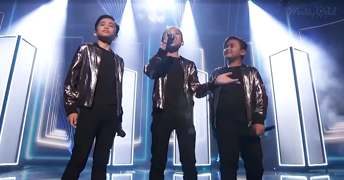 Three children in shiny jackets perform on stage under bright lights and geometric shapes.