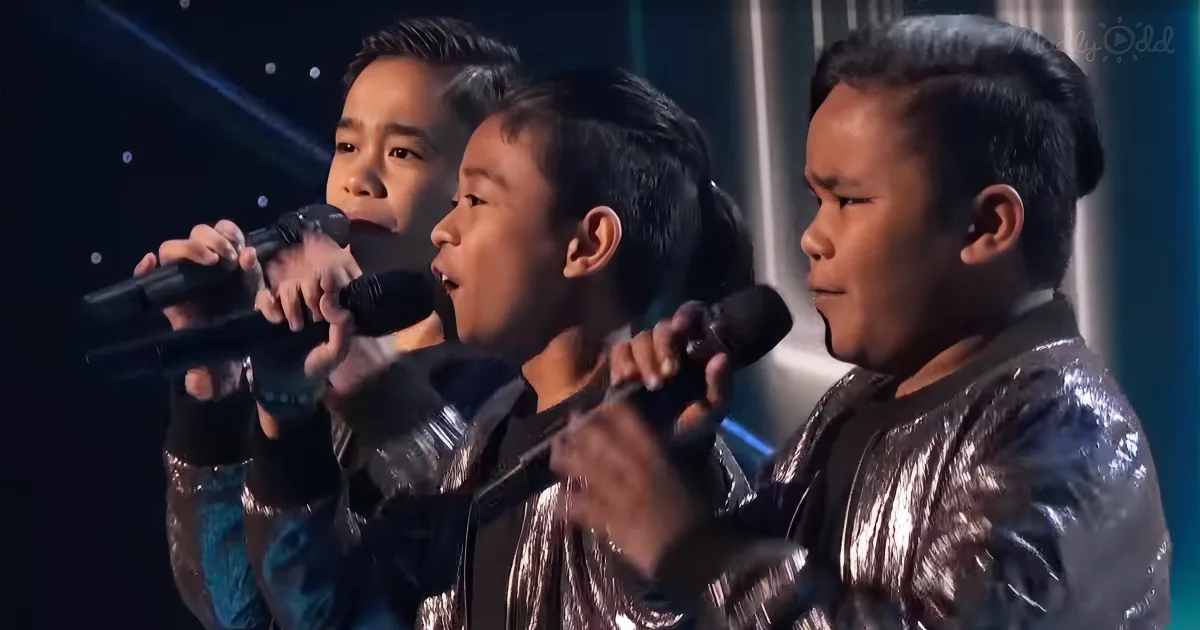 Three individuals in shiny jackets sing into microphones on a stage with a dark background.