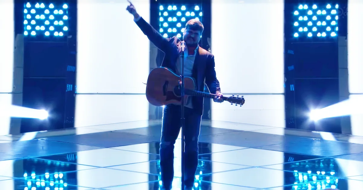 A man in a suit plays an acoustic guitar on a reflective stage with bright lights.