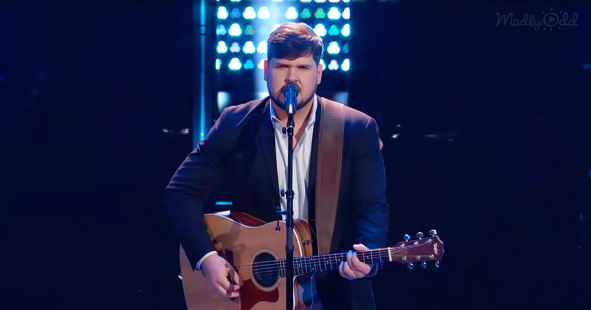 A man in a suit sings into a microphone while playing an acoustic guitar on stage.