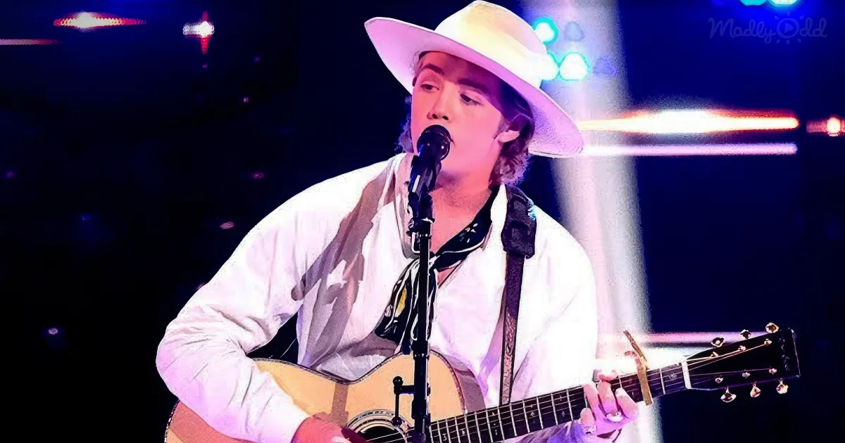 A man in a white shirt and hat plays an acoustic guitar on stage, singing into a microphone.