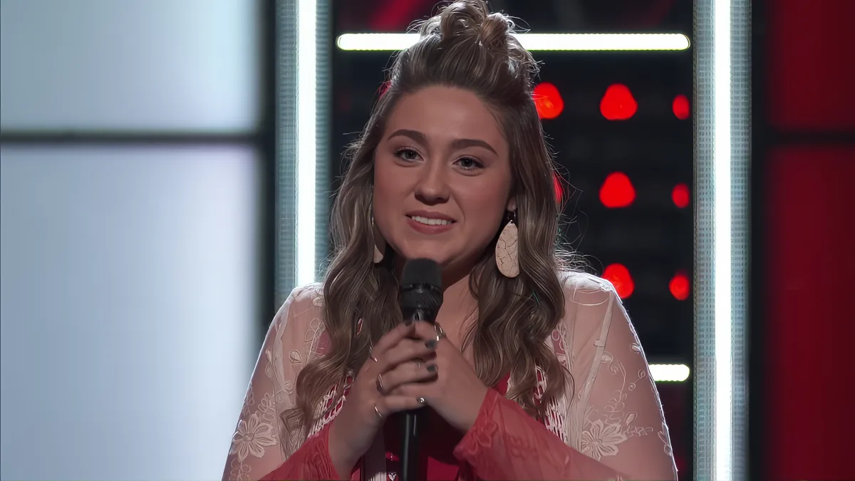 A woman with long, wavy hair in a bun, wearing a red and white lace top, smiles while holding a microphone on stage.