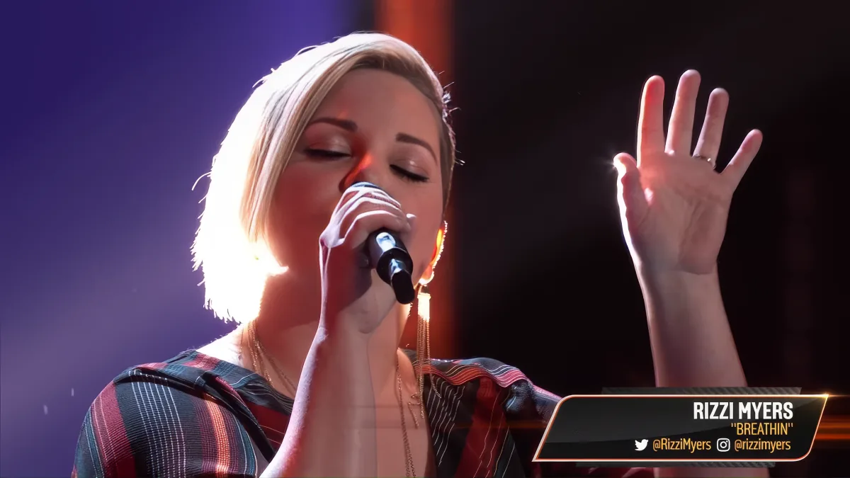 A woman sings passionately, holding a microphone, with her hand raised, illuminated by stage lights.