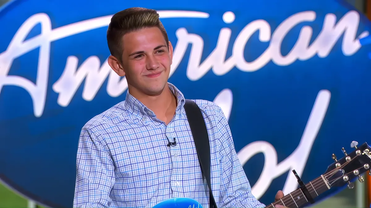 A young man in a blue checkered shirt holds a guitar, smiling in front of an "American Idol" backdrop.