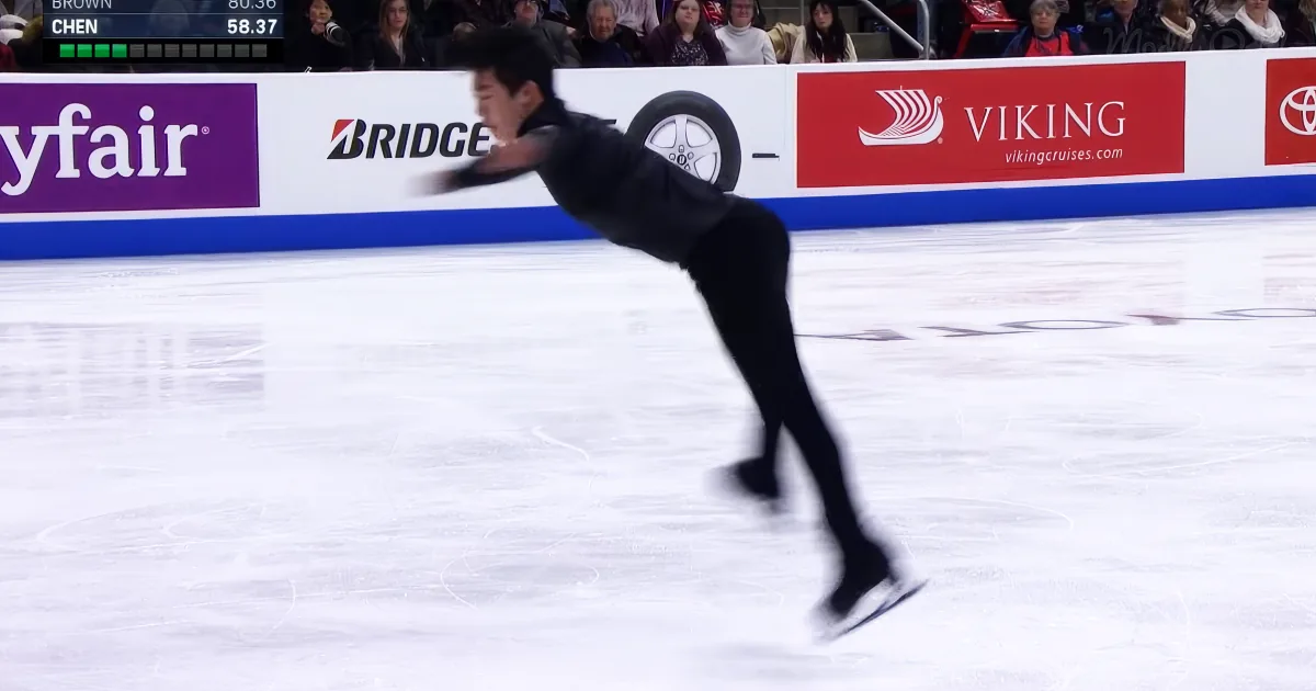 A figure skater in a black outfit glides on ice, with blurred motion and spectators in the background.