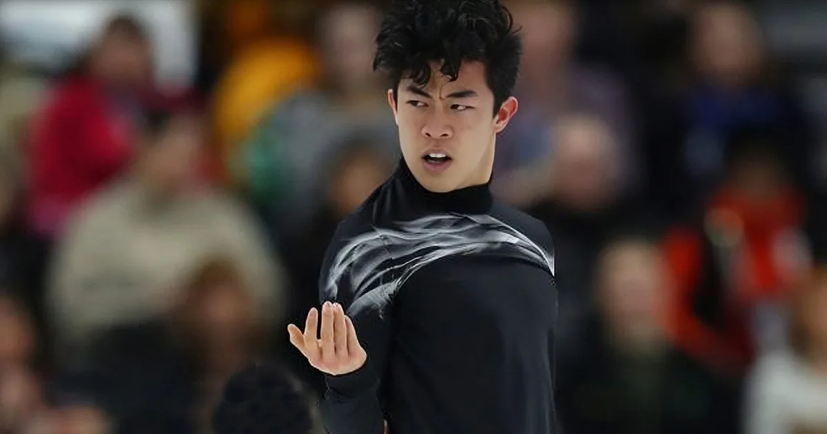 A figure skater in a black outfit with a white design gestures with his right hand while performing on ice.