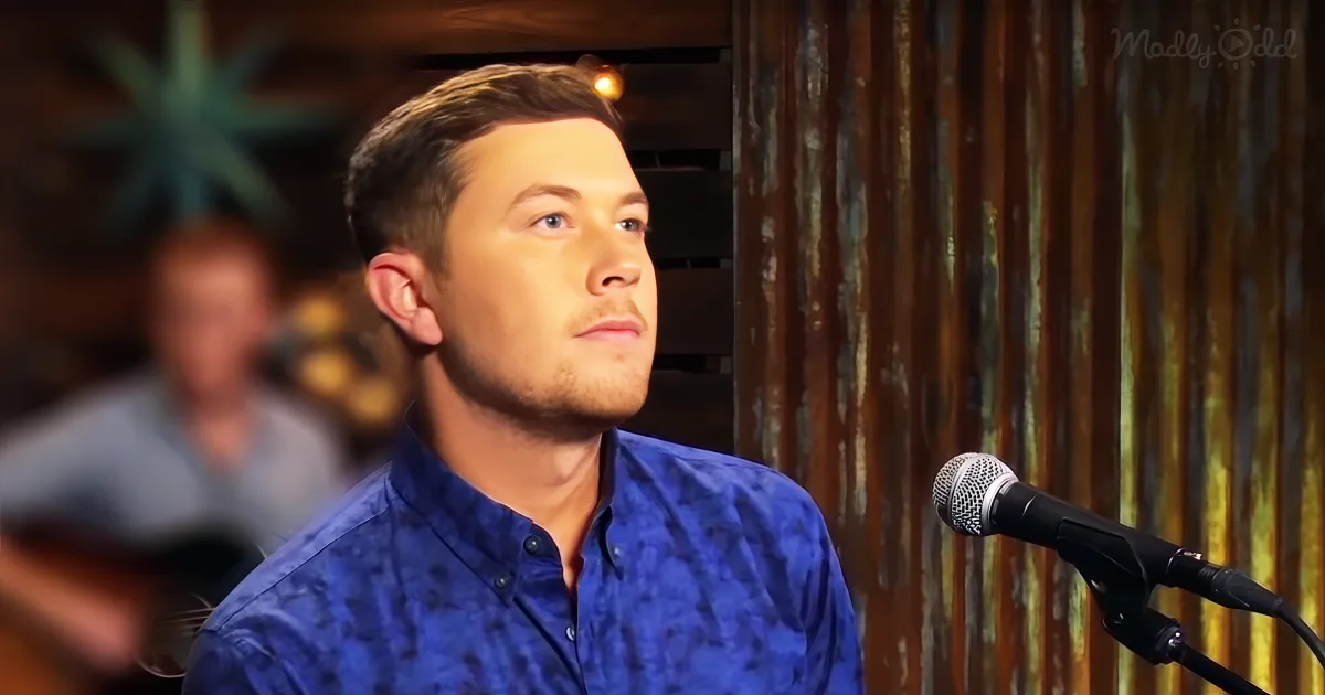 A man in a blue shirt sings into a microphone in a dimly lit room with a wooden backdrop.