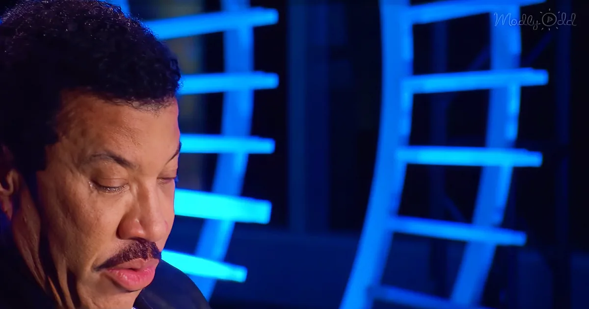 A man with a mustache and closed eyes, wearing a dark suit, is in front of a blue-lit background with geometric shapes.