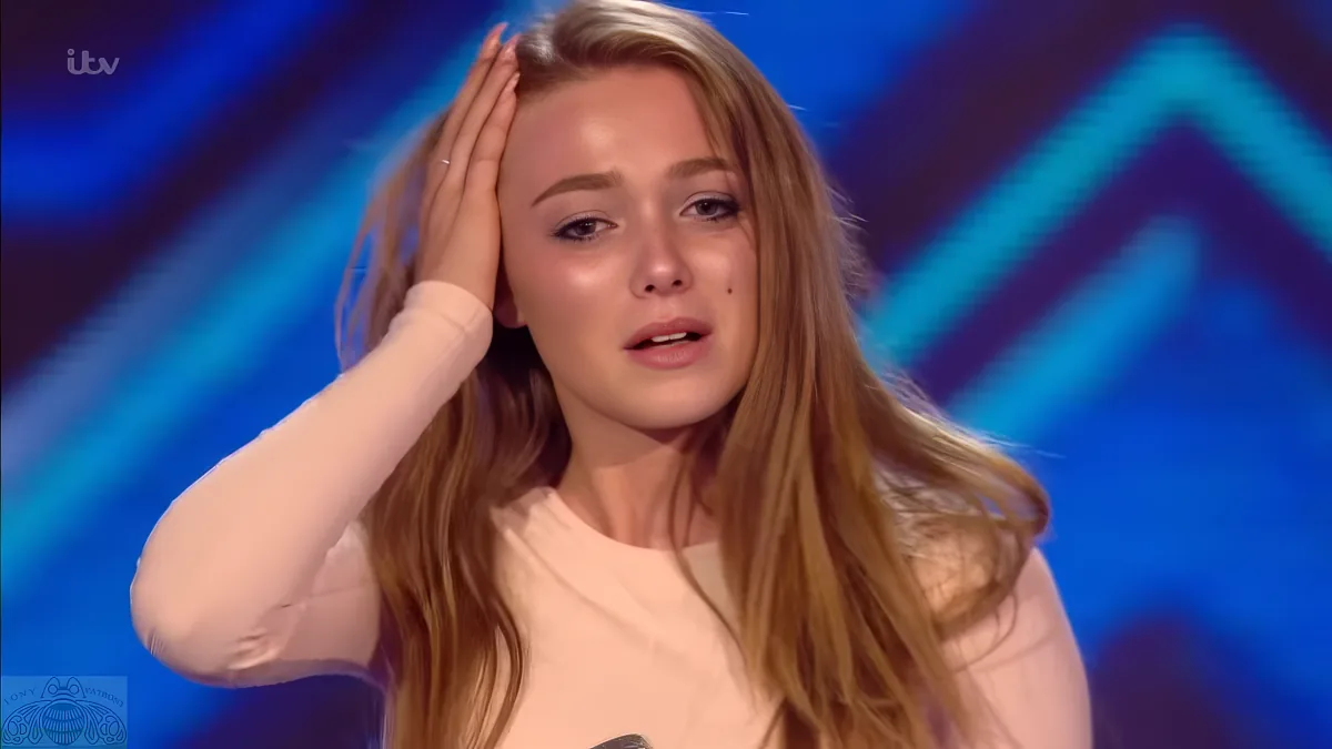 A person with long blonde hair, wearing a white top, is holding their hand to their head, appearing emotional.