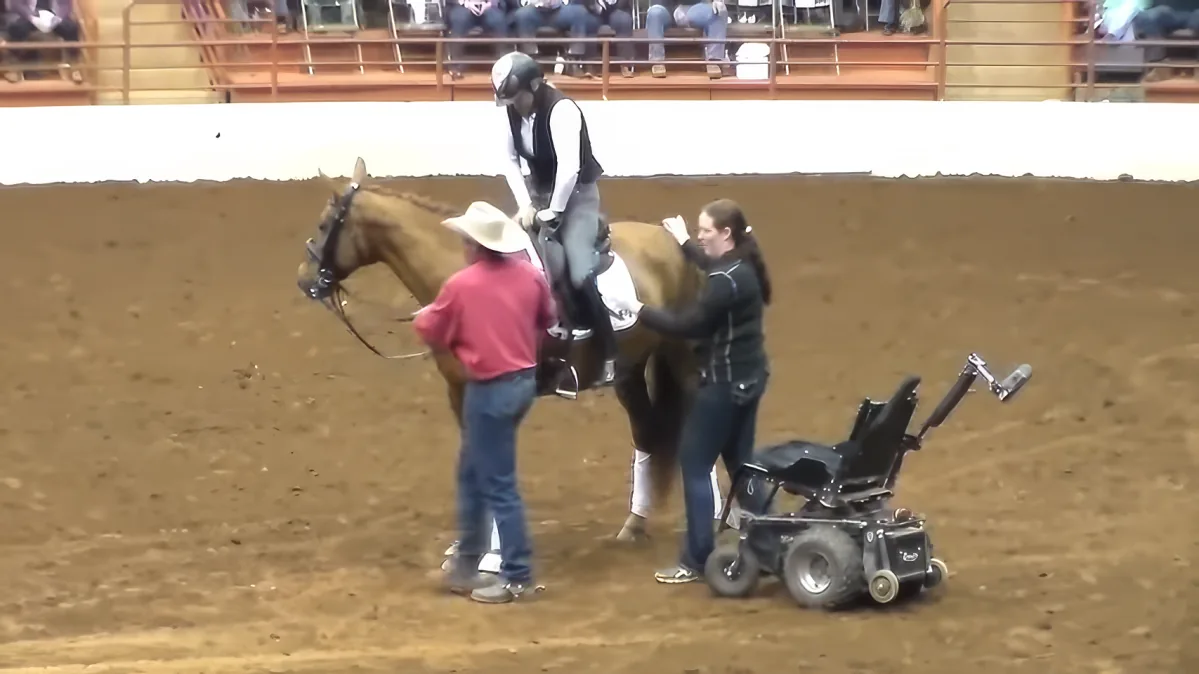 A person on a horse is being assisted by two others near a power wheelchair in an indoor arena.