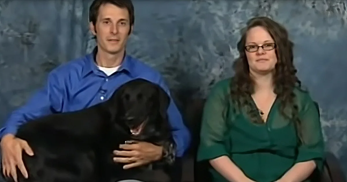 A man in a blue shirt holds a black dog, while a woman in a green shirt sits beside him against a blue background.