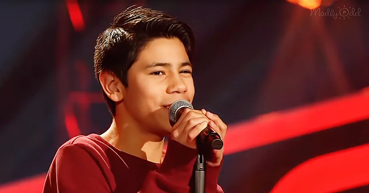 A young person in a red shirt sings into a microphone on stage with red lighting.