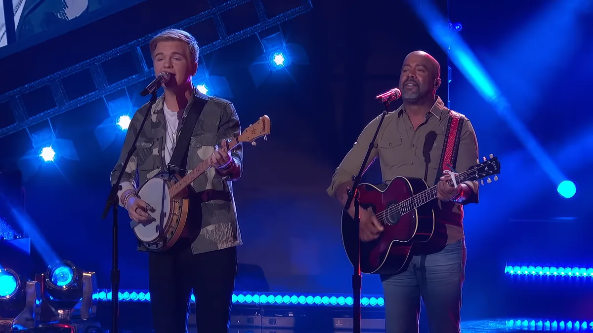 Two musicians perform on stage, one playing banjo, the other guitar, under blue lights.