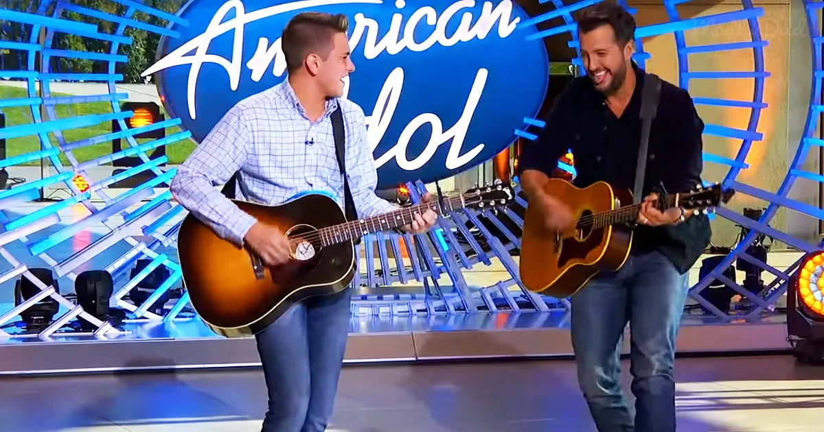 Two men play acoustic guitars on stage with "American Idol" backdrop.