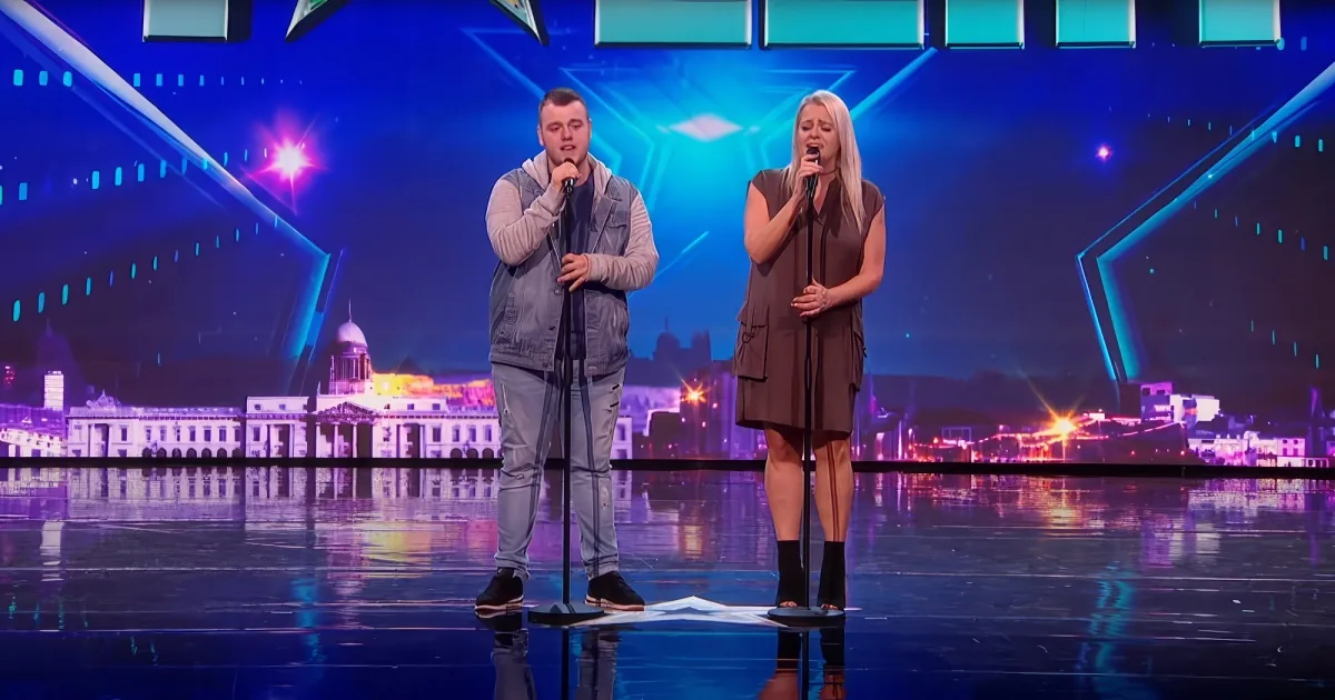 A man and woman sing on a stage with a cityscape backdrop and colorful lighting.
