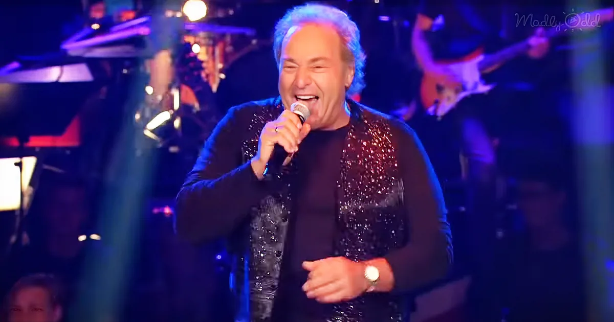 A man in a glittery jacket sings into a microphone on stage with a band in the background.