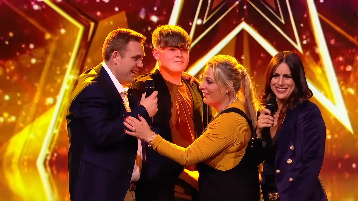 A man in a suit holds a microphone, surrounded by three smiling people on a stage with bright lights.