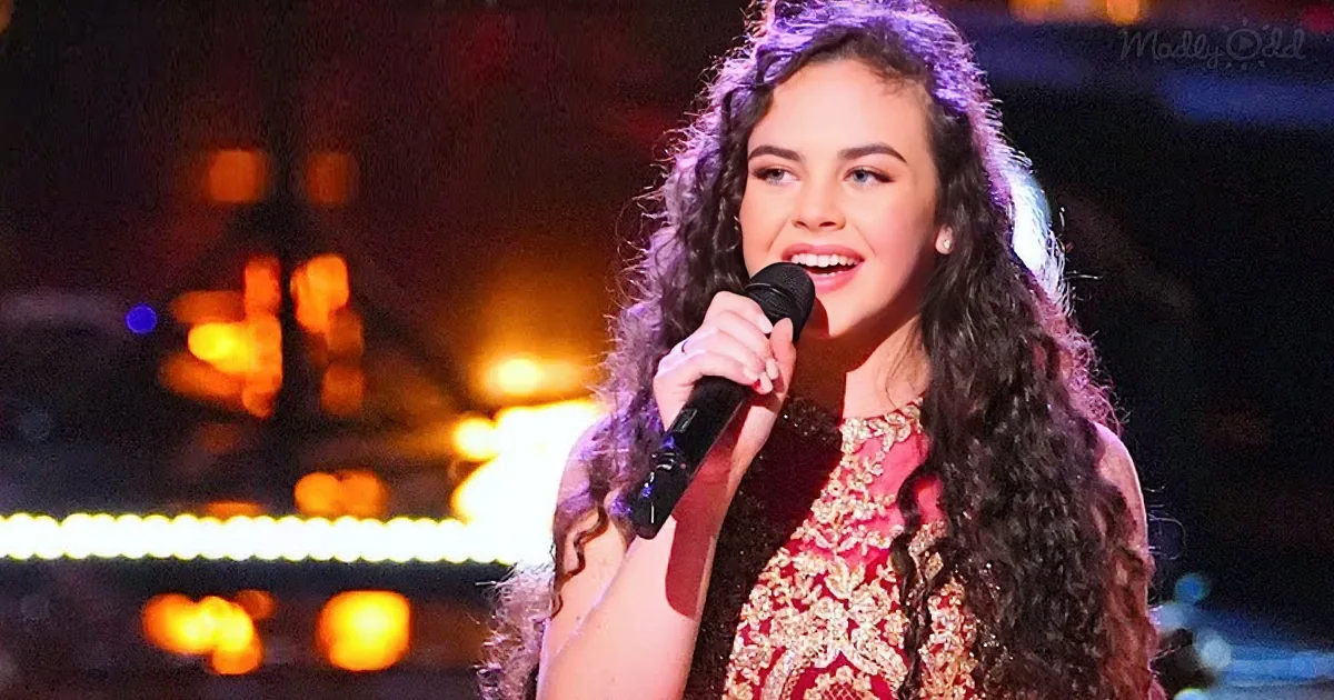 A woman with long, curly hair holds a microphone, wearing a red and gold dress, singing on stage with warm lights in the...
