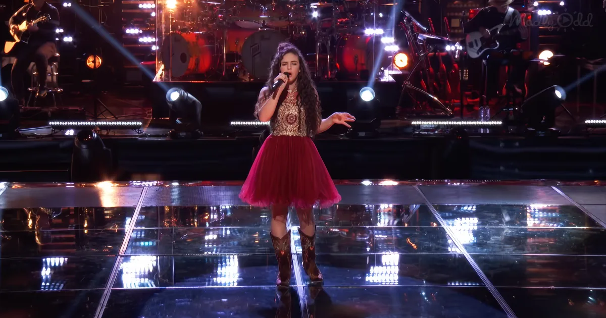 A woman in a red dress and cowboy boots sings on stage with a band and bright lights.