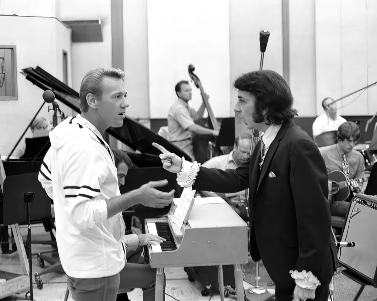 A man in a white shirt points while another in a suit listens, surrounded by musicians in a recording studio.
