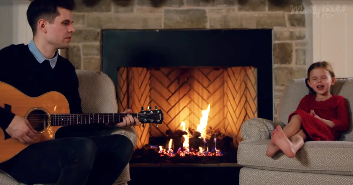 A man plays guitar while a child sits on a couch near a lit fireplace.