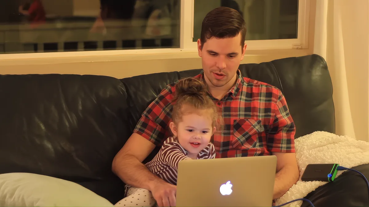 A man in a red plaid shirt sits on a black couch with a child, using a laptop.