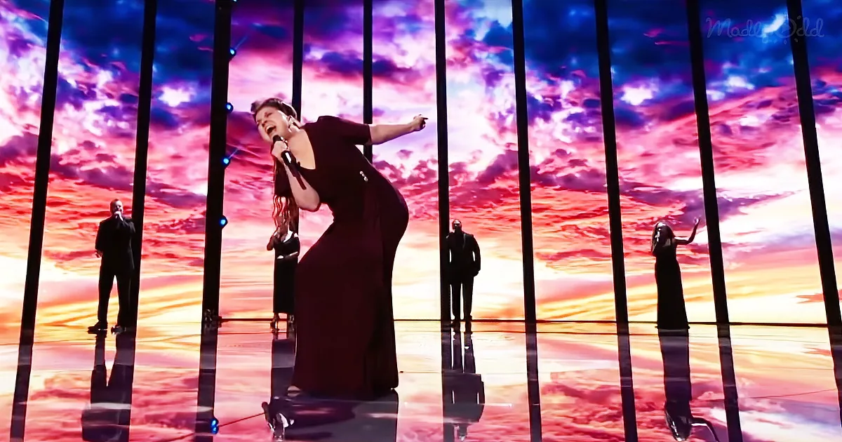 A woman in a black dress sings passionately on stage with a vibrant sunset backdrop and reflective pillars.