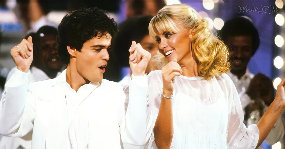 A man and woman in white outfits dance enthusiastically, surrounded by a crowd, with bright lights in the background.