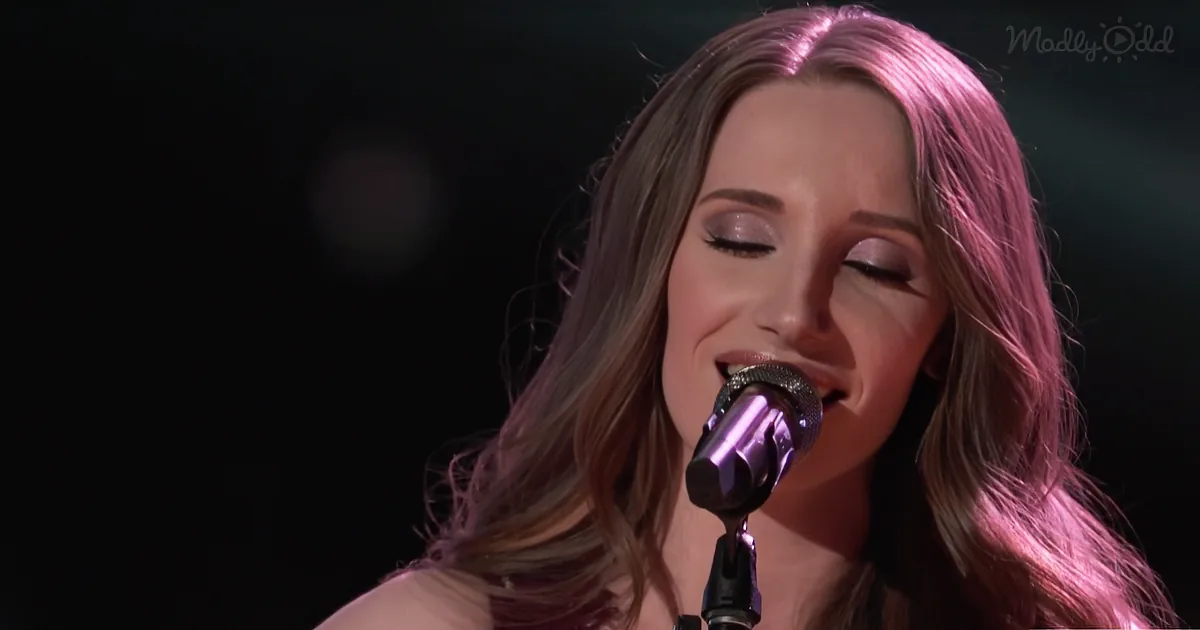A woman with long, wavy hair sings into a microphone, her eyes closed, illuminated by purple stage lighting.