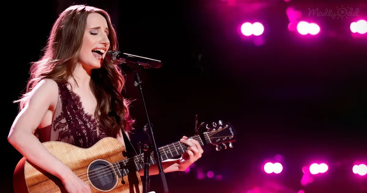 A woman sings passionately, playing an acoustic guitar on stage with vibrant pink lights.