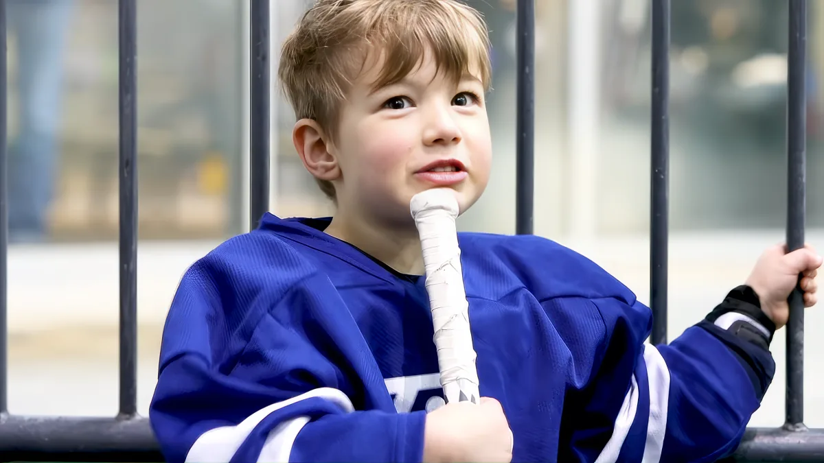 A young boy in a blue hockey jersey holds a hockey stick, looking up with a thoughtful expression.
