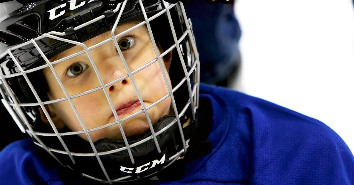 A young hockey player in a blue jersey and black helmet with a cage, looking intently to the side.