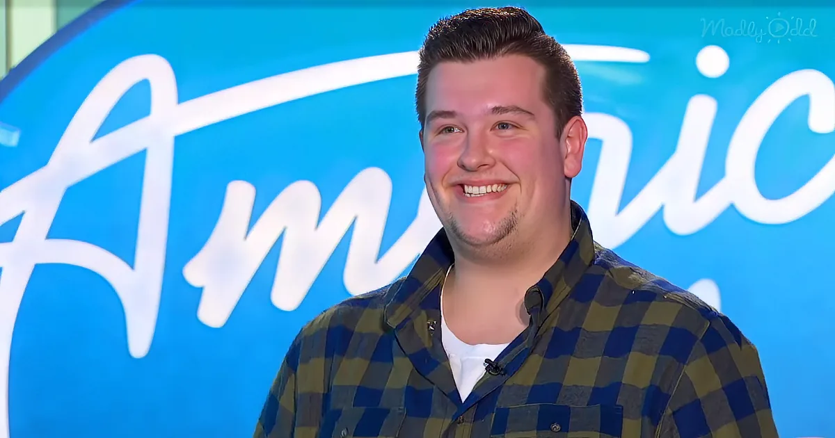 A smiling man in a plaid shirt stands in front of a blue "America's Got Talent" backdrop.