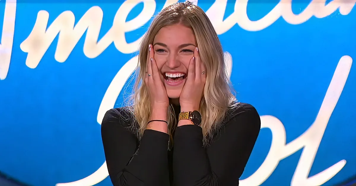 A woman with blonde hair, wearing a black top and a gold watch, smiles widely with her hands on her cheeks.