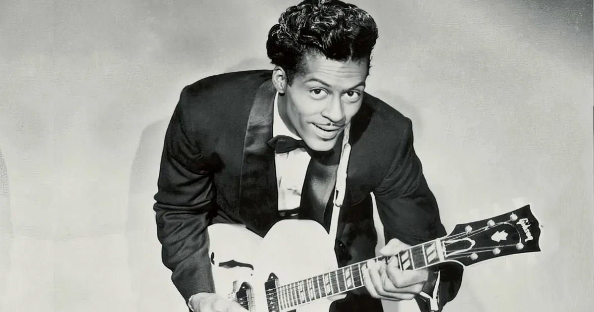 A man in a tuxedo plays a white guitar, smiling and looking up.