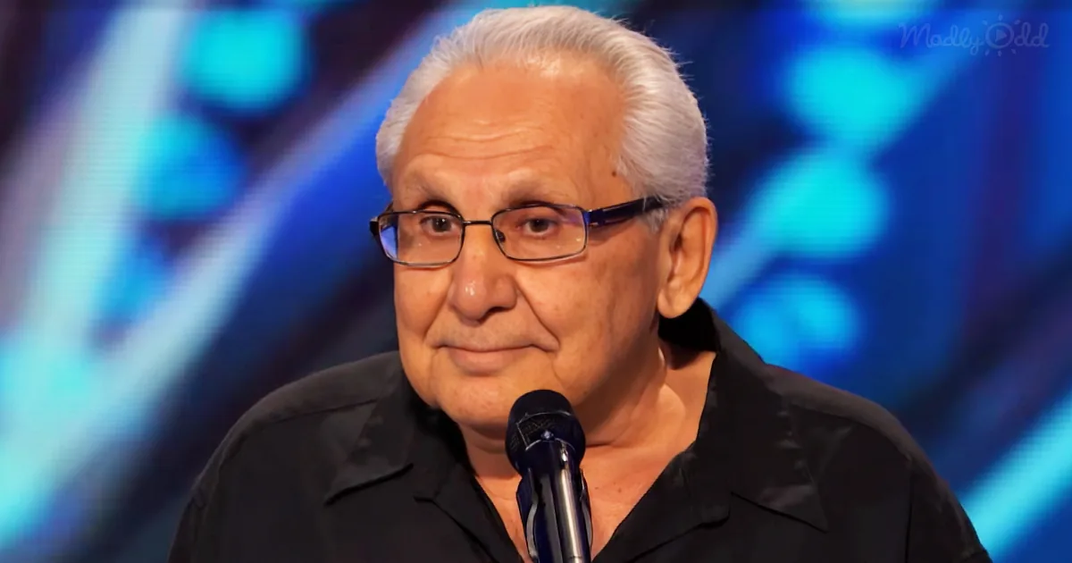 An older man with white hair and glasses, wearing a black shirt, speaks into a microphone against a blue-lit background.