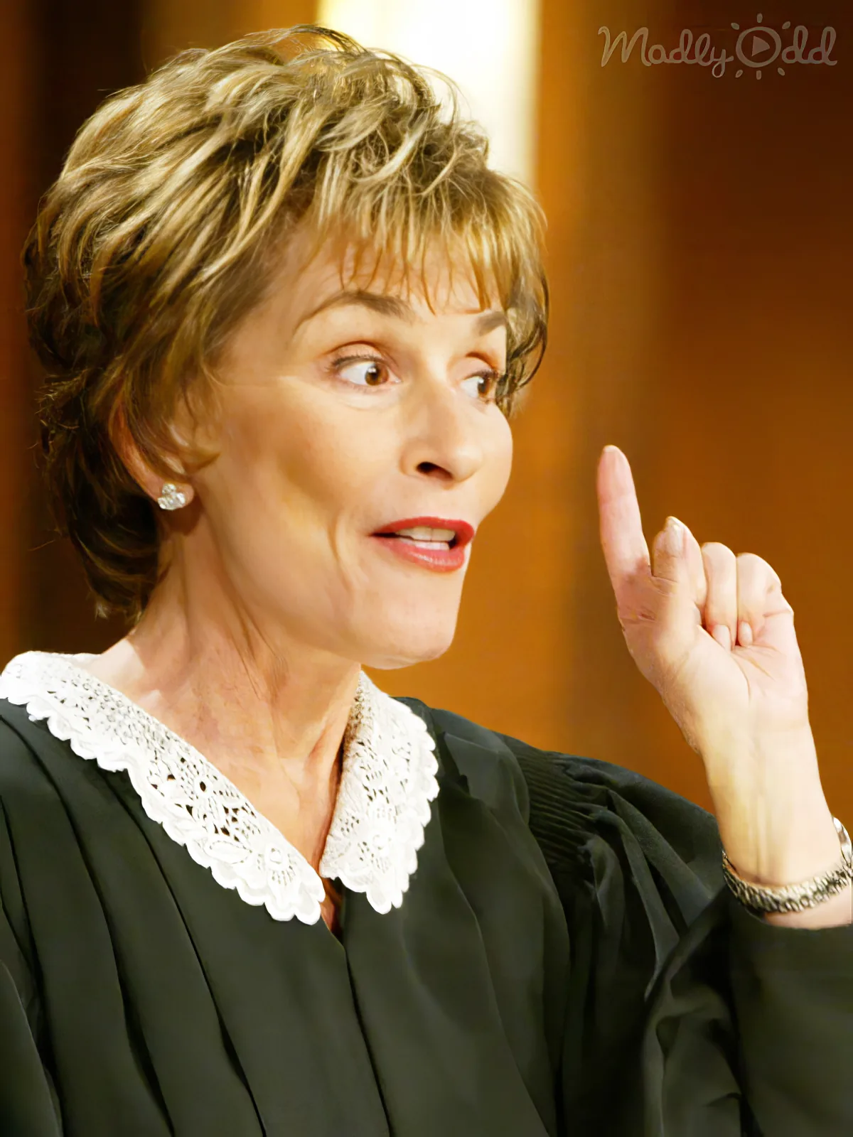 A woman in a black robe with a white lace collar gestures with her right hand while looking to the side.