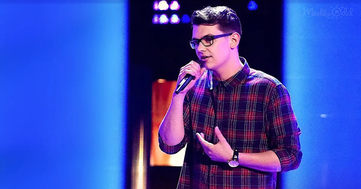 A man in a plaid shirt holds a microphone, gesturing with his hands on stage.