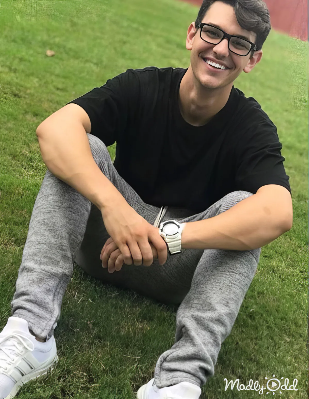 A person in a black shirt and gray sweatpants sits on grass, smiling, with a watch on their wrist.