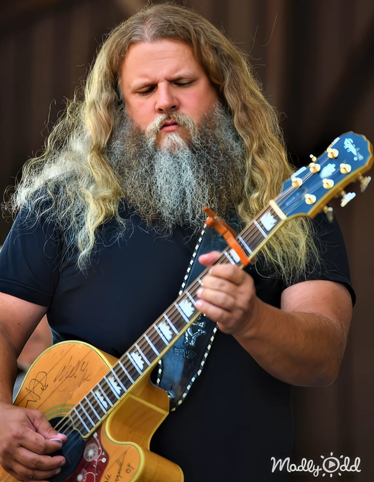 A man with long hair and a beard plays a guitar with a distinctive headstock design.