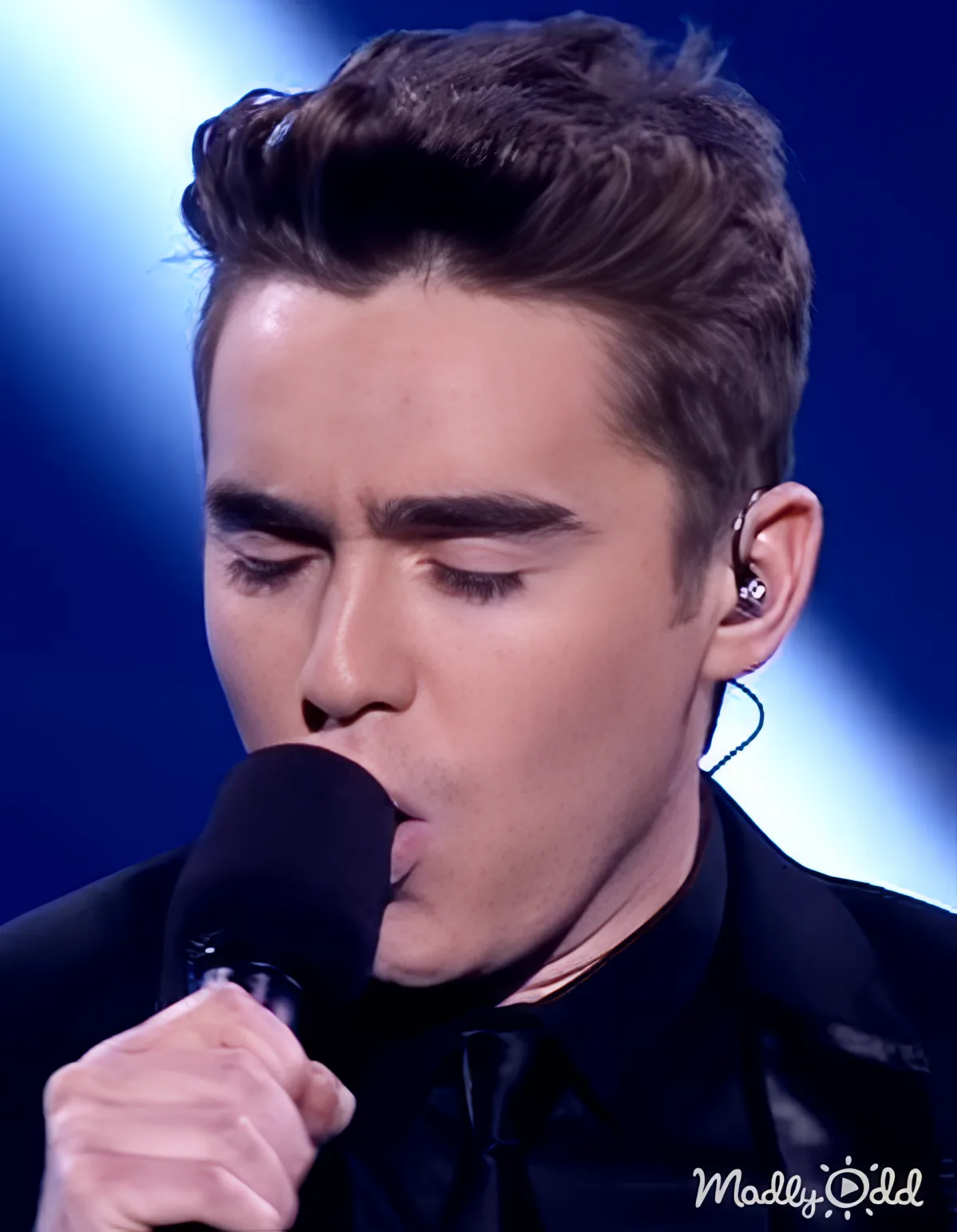 A man in a black shirt and tie sings into a microphone, wearing an earpiece, with a blue background.