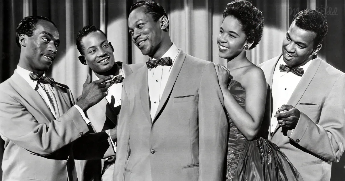 A man in a suit is being adjusted by another man, while a woman and another man look on.