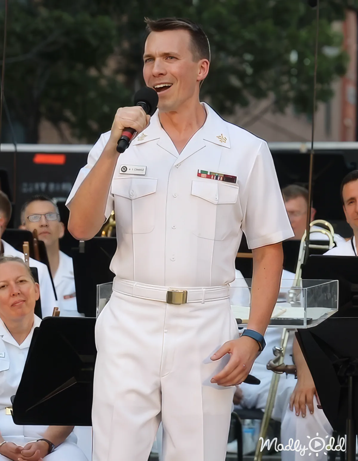A man in a white naval uniform speaks into a microphone, smiling, with a band in the background.
