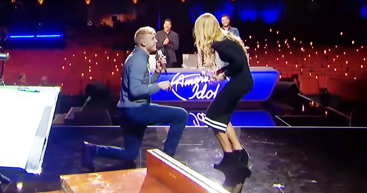 A man kneels on stage, holding a microphone, proposing to a woman in a black dress.