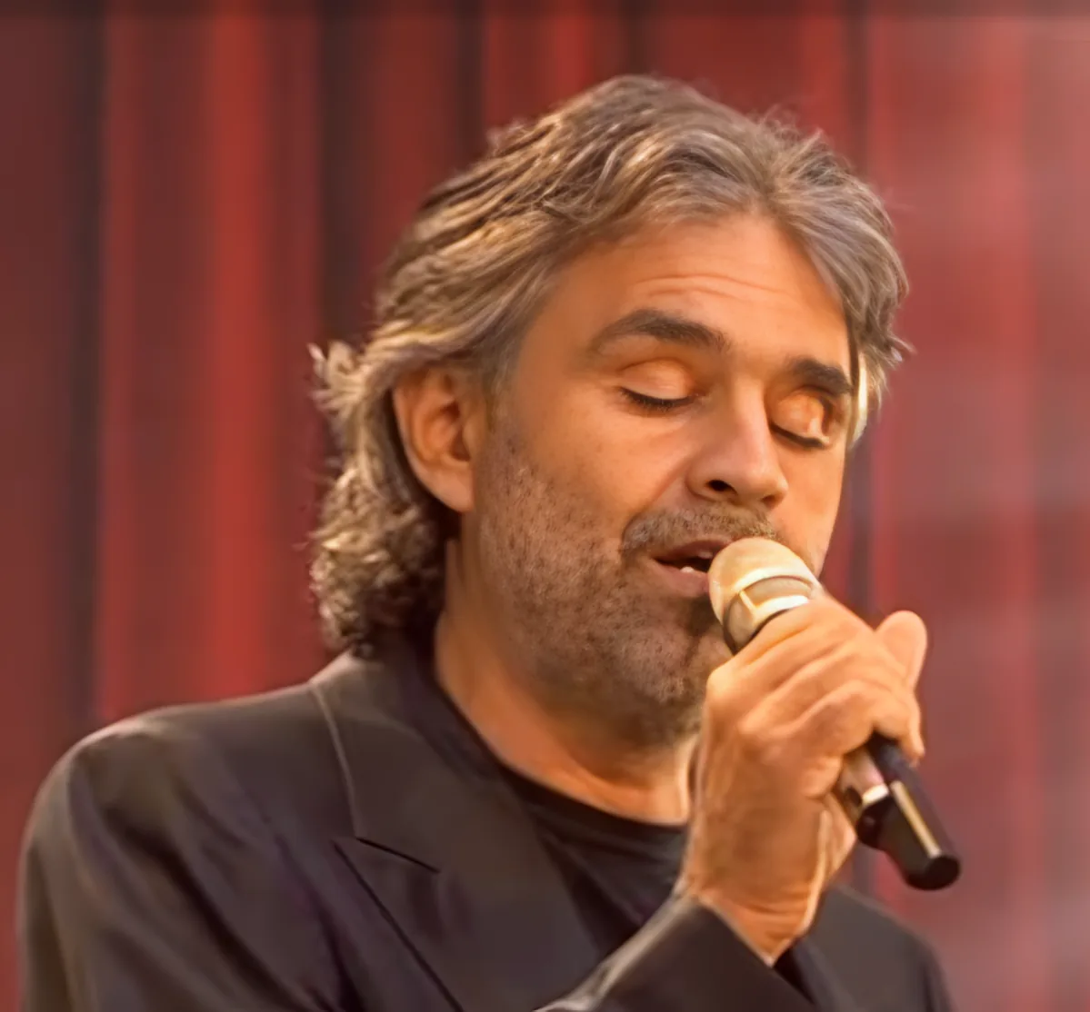 A man with long hair and a beard sings into a microphone, wearing a black shirt and blazer.